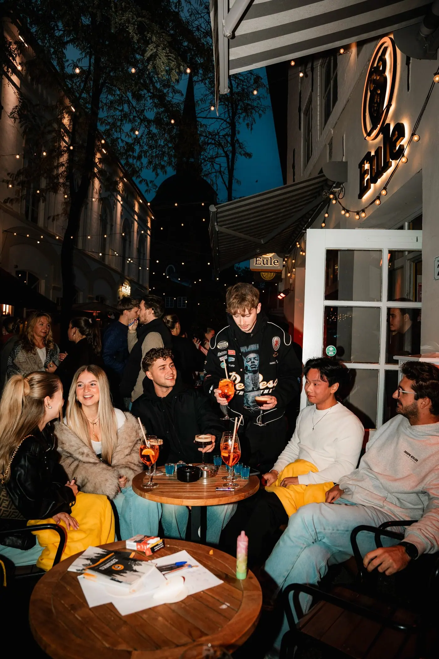 Gäste sitzen abends draußen an Tischen unter dem leuchtenden Eule-Schild